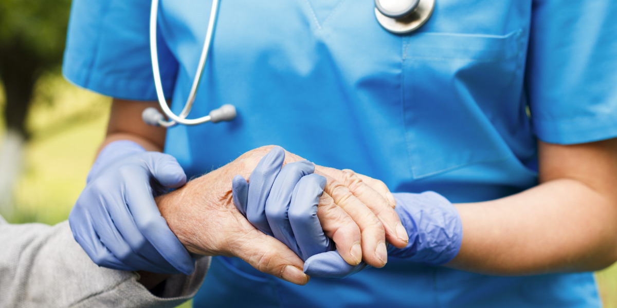 Nurse wearing stethescope and gloves holding an elderly persons hand.