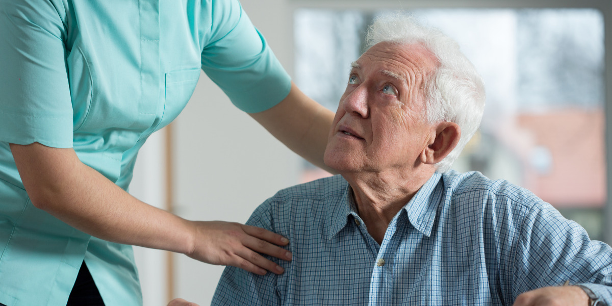 Nurse comforting an elderly man wearing a blue chequered shirt.