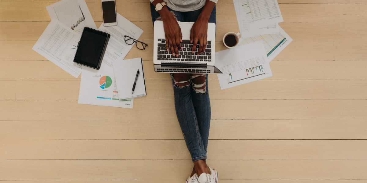 Person sat on the floor surrounded by paperwork working on laptop.