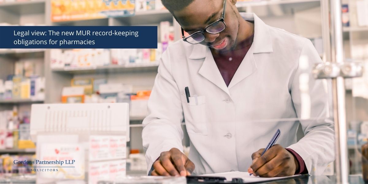 Man with a doctor jacket on writing on a clipboard with medicine cabinets in the background.