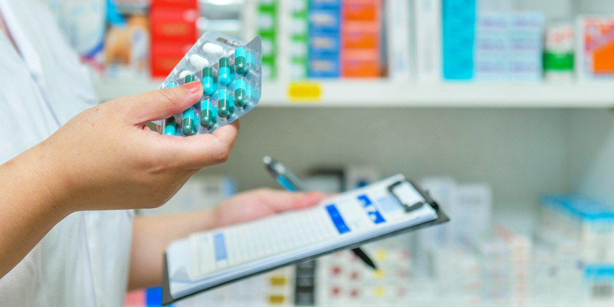 Someone holding pills and a clipboard with a medicine shelf in the background.