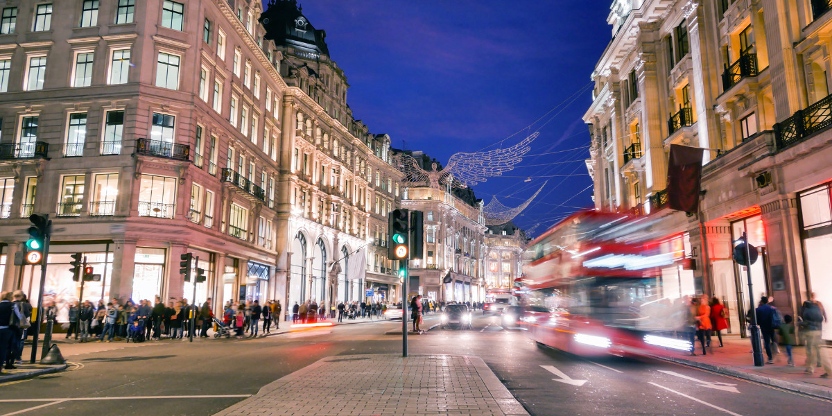 A bustling city street at twilight with illuminated buildings, a blur of a double-decker bus in motion, and pedestrians waiting to cross, under a canopy of decorative lights.