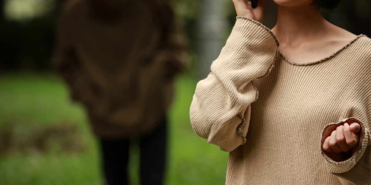 A person in the foreground wearing a beige sweater with a focus on the sweater's texture, while another individual is slightly blurred in the background amidst a green, natural setting.