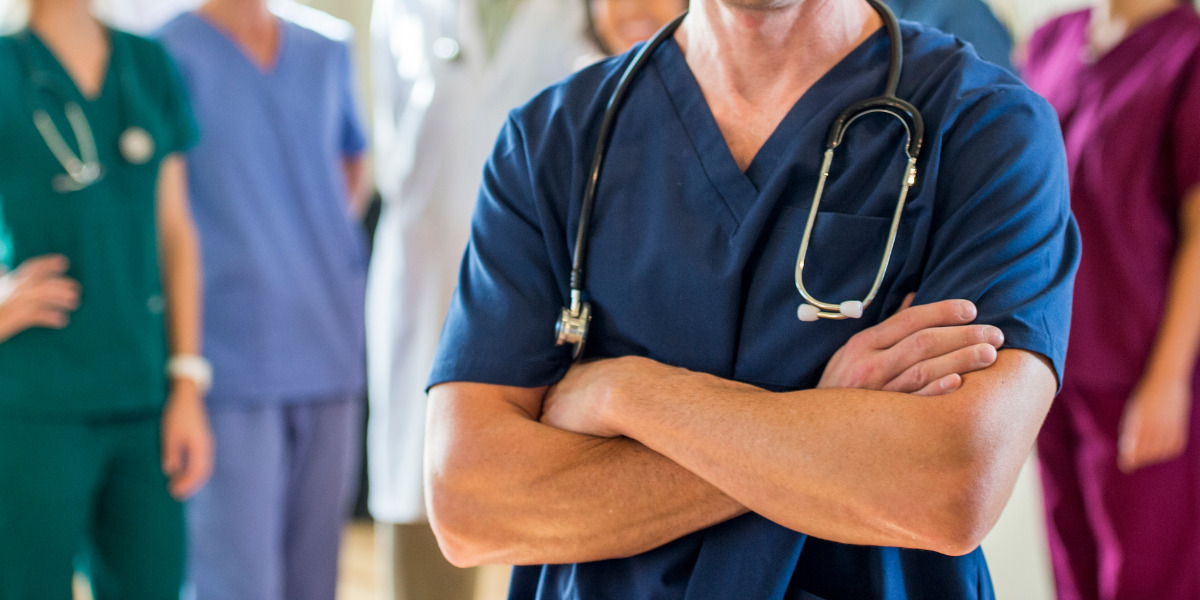 A confident healthcare professional stands with arms crossed, a stethoscope draped around the neck, as colleagues gather in the blurry background, depicting a scene of medical teamwork and readiness.