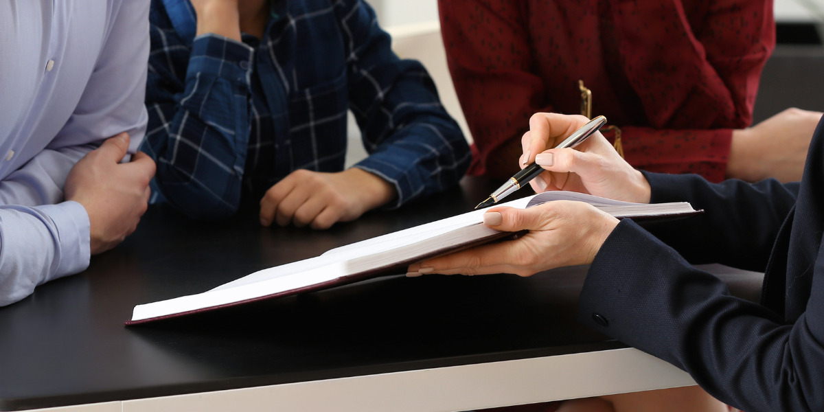 Four individuals at a meeting, with focus on the hands of one person pointing to a document, suggesting a discussion or review of its contents.