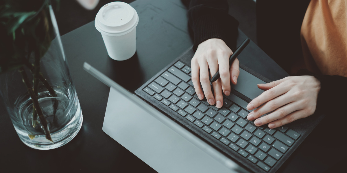 A person working on a tablet with a keyboard attachment in a modern, minimalist setting with a paper cup of coffee nearby.