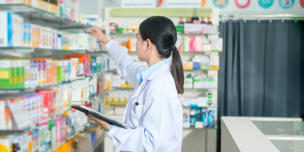A pharmacist in a white coat checking inventory in a pharmacy, with shelves stocked full of medicine boxes.
