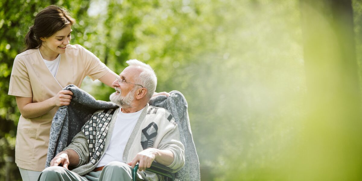 A woman helps an elderly man in a wheelchair, reflecting the commitment to sustainable practices in care facilities.