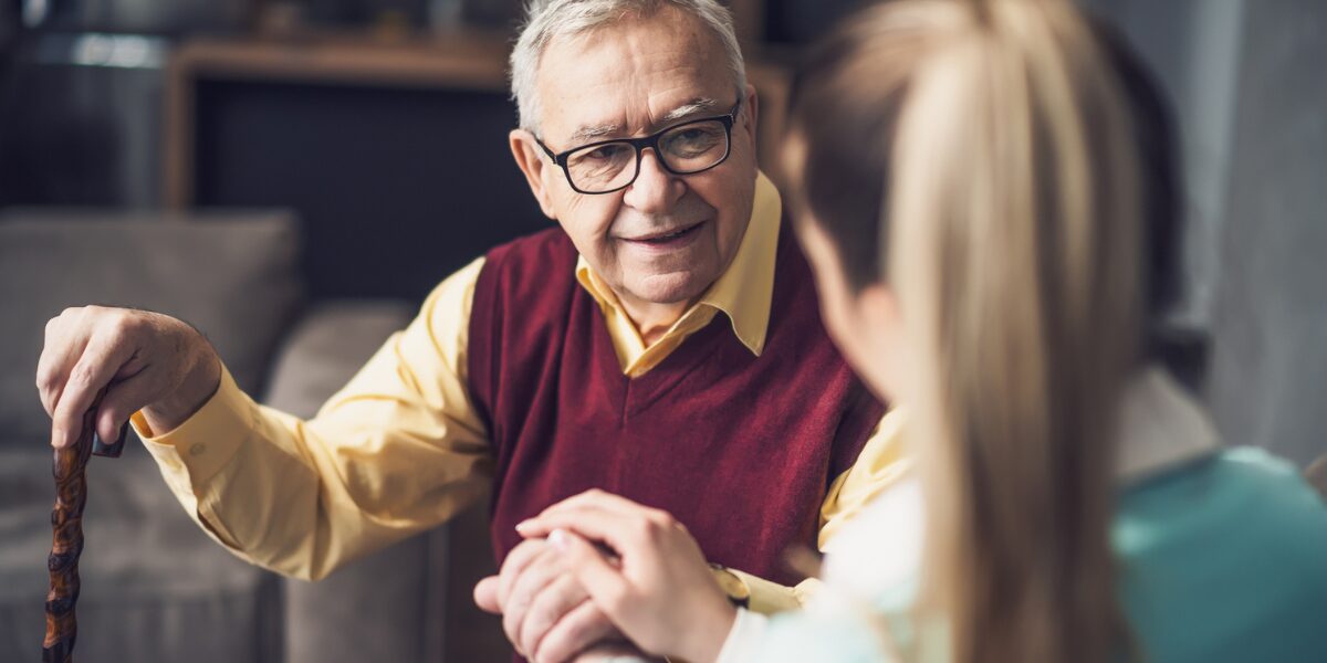 An older gentleman with glasses and a cane is engaged in discussion with a young female carer, reflecting a warm conversation, portraying the importance of genuine care,