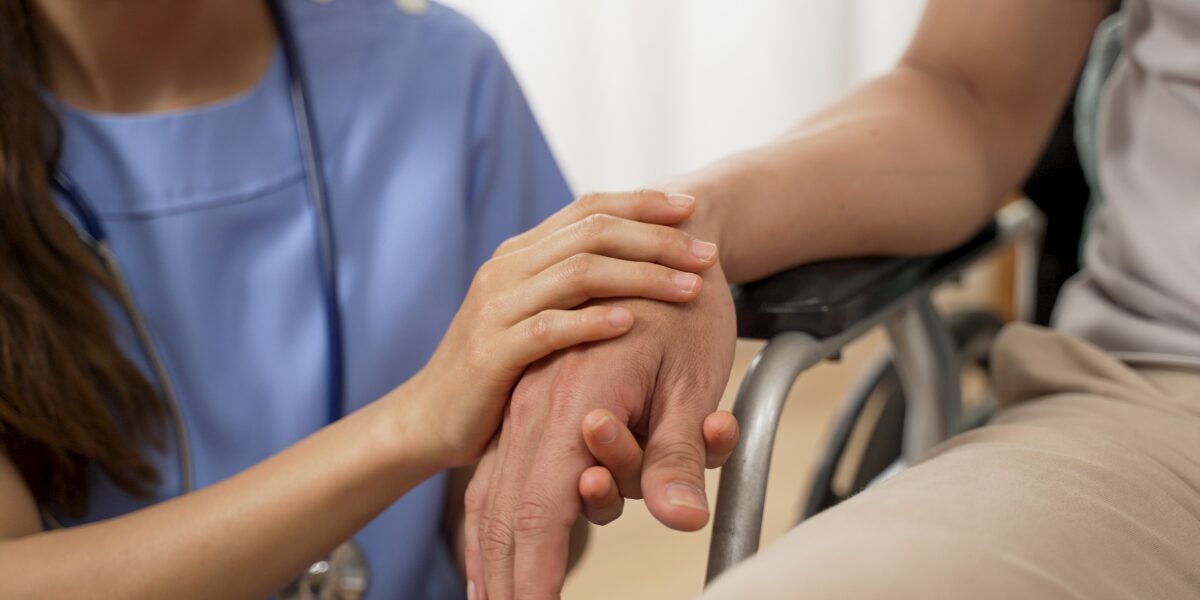 A nurse gently holds a wheelcheair bound patient's hand, providing comfort and support in a domiciliary care setting.